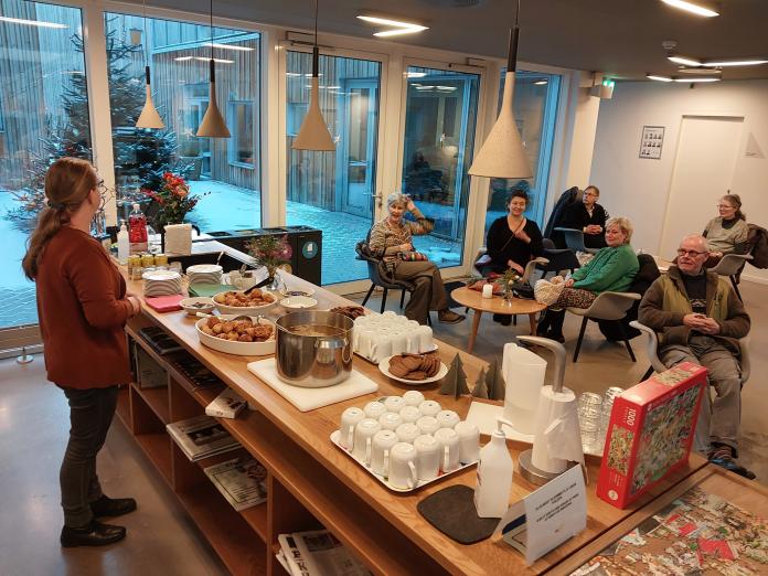 Kvinde ved bord i caféen i Center for Kræft og Sundhed med gløgg og æbleskriver. Der sidder flere mennesker i stole bagved og kigger på.